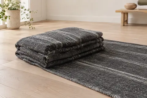 Couverture de yoga Manduka gris foncé à rayures, pliée sur un sol en bois clair.