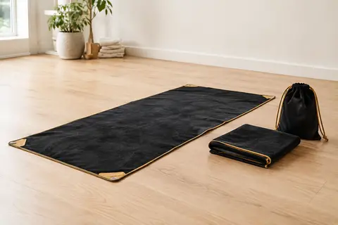 Serviette de yoga noire et dorée déployée, avec son sac à cordon posé à côté sur un sol en bois clair